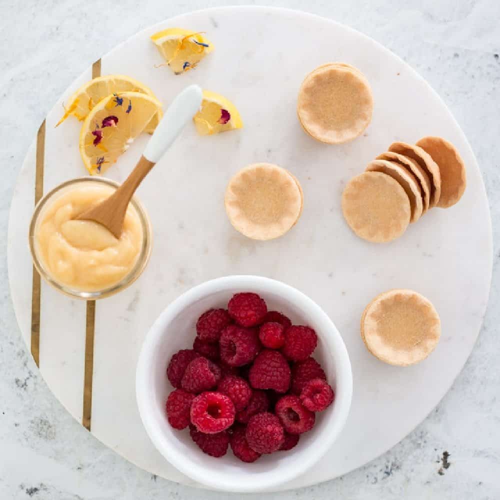 Overhead view of lemon tarts with a bowl of raspberries on a white plate.