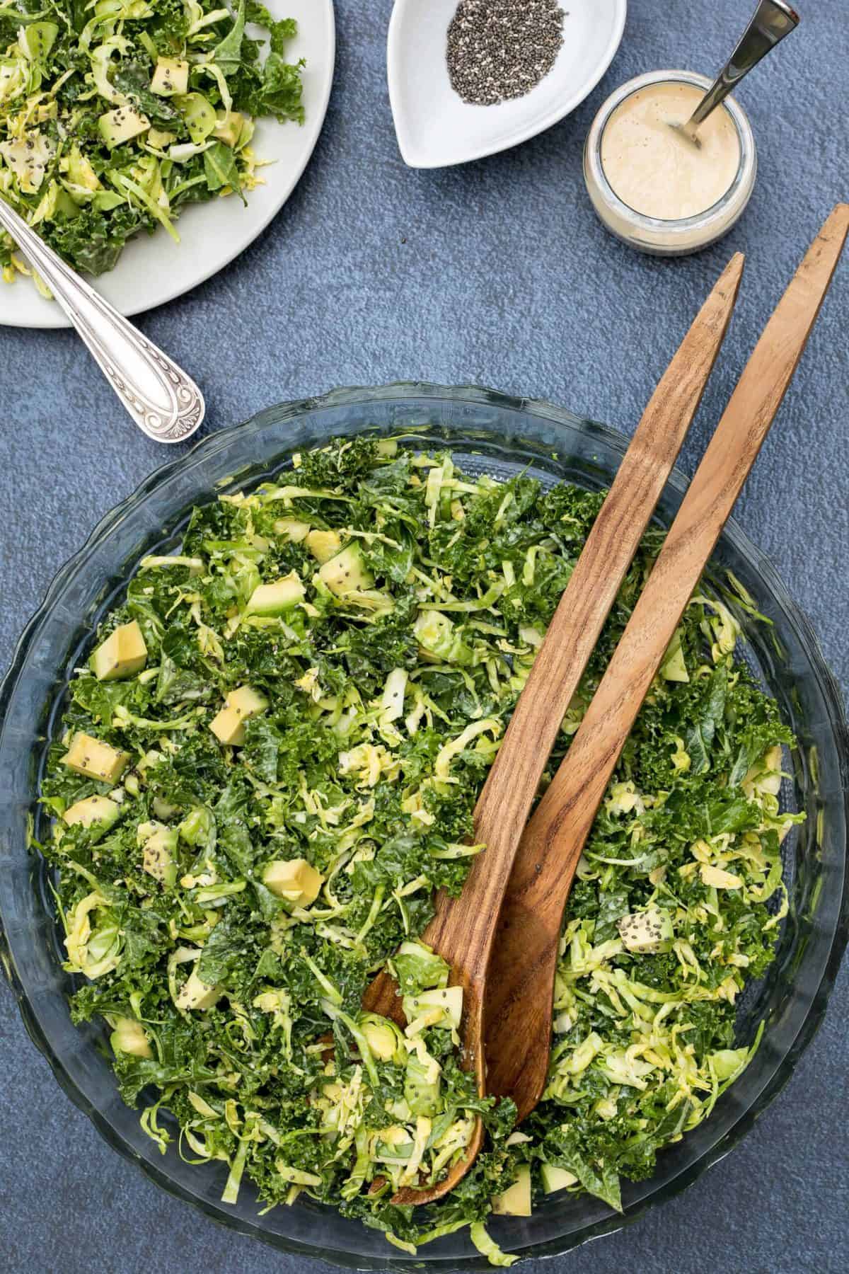 Overhead view of a bowl full of kale and Brussels sprouts salad with miso-ginger dressing, a wooden serving spoon, and a small plate of salad.