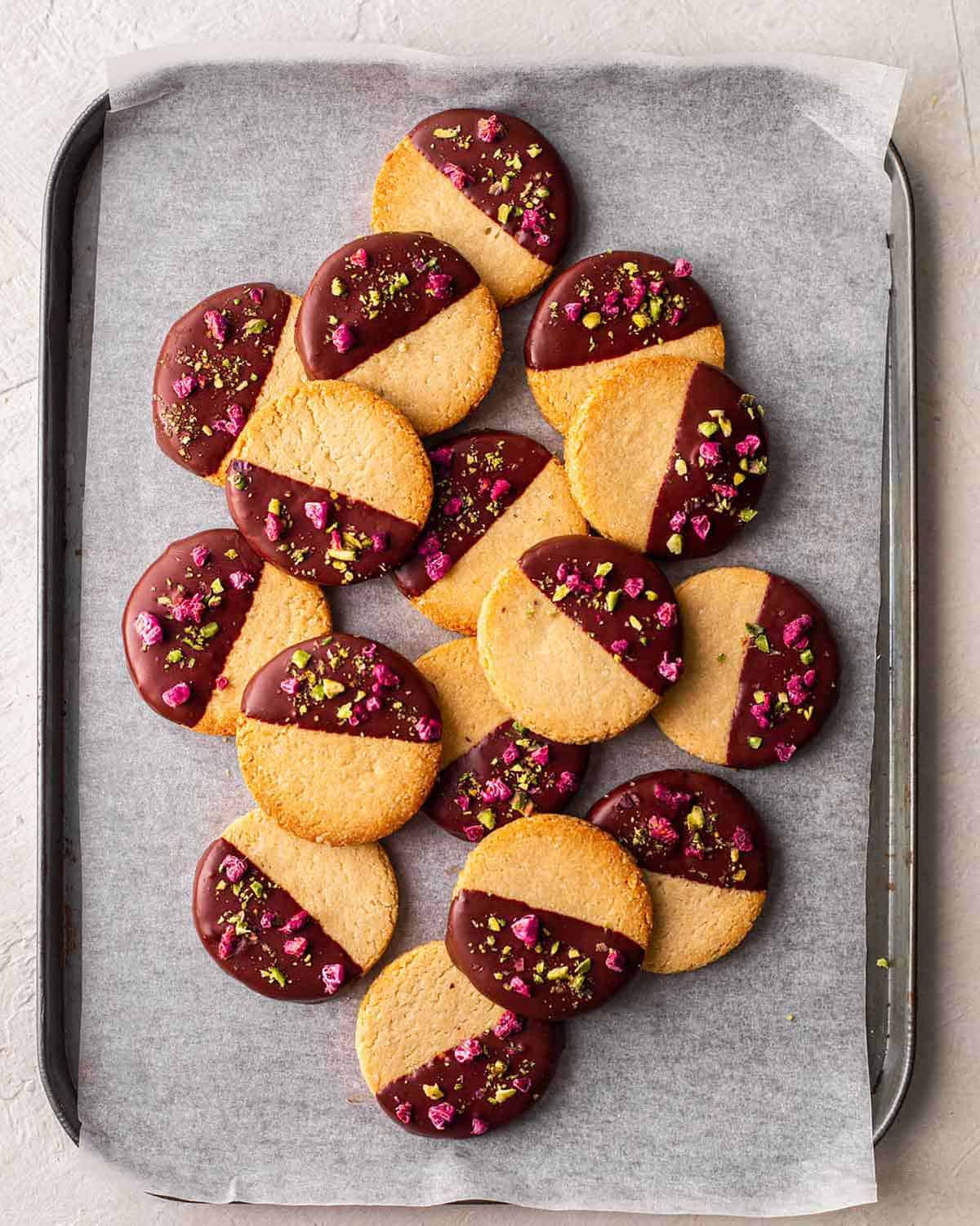 An overhead view of Vegan Almond Flour Shortbread Cookies dipped in chocolate, topped with pistachios and freeze-dried raspberries, placed in a baking tray.
