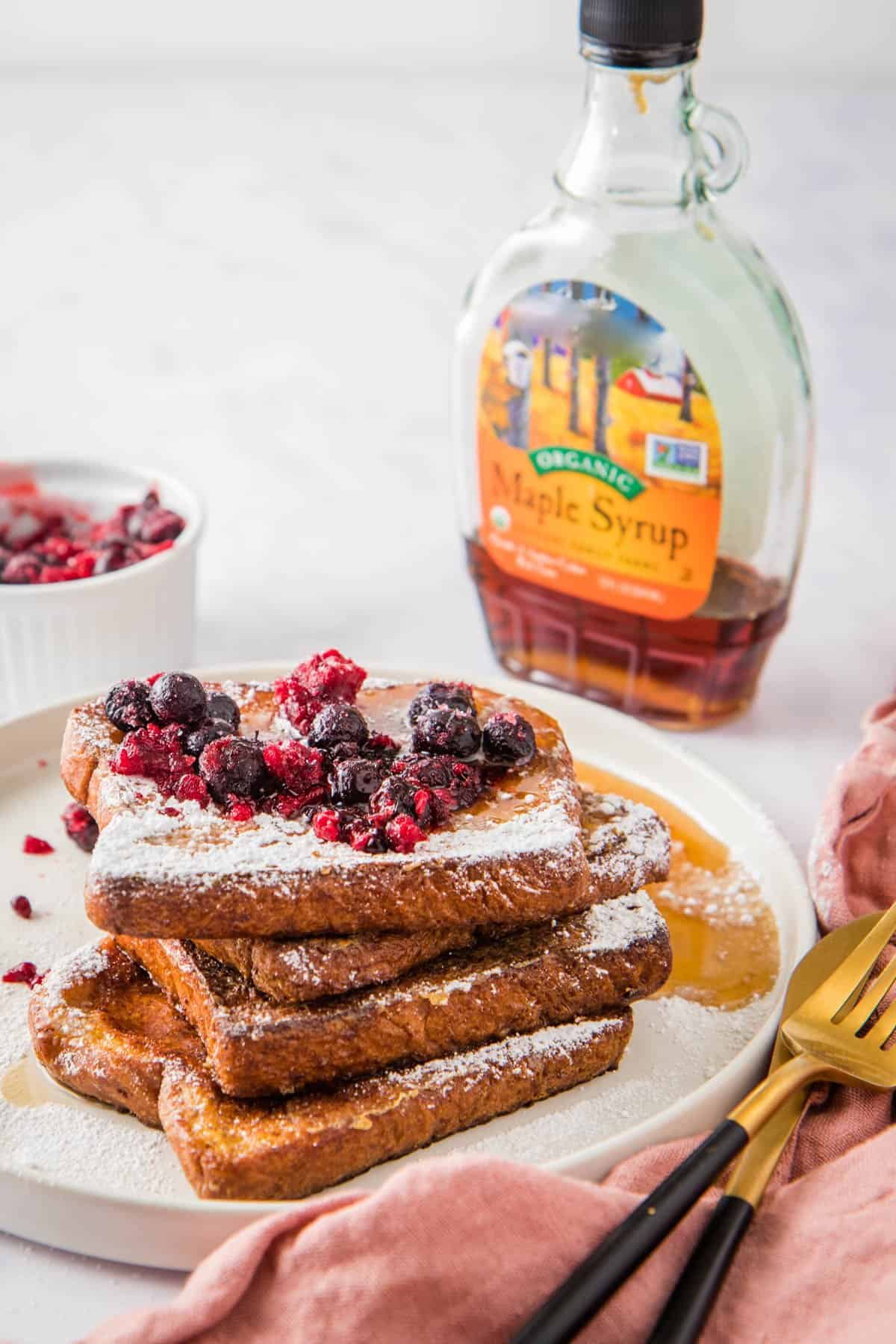 Close up of stack of French toast with berries and powdered sugar on white plate, maple syrup and extra berries in the background