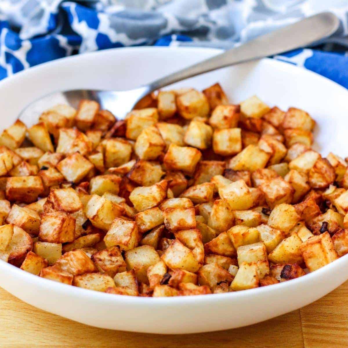 A close-up view of air-fried home fries in a white bowl and has a blue and white napkin in the background.