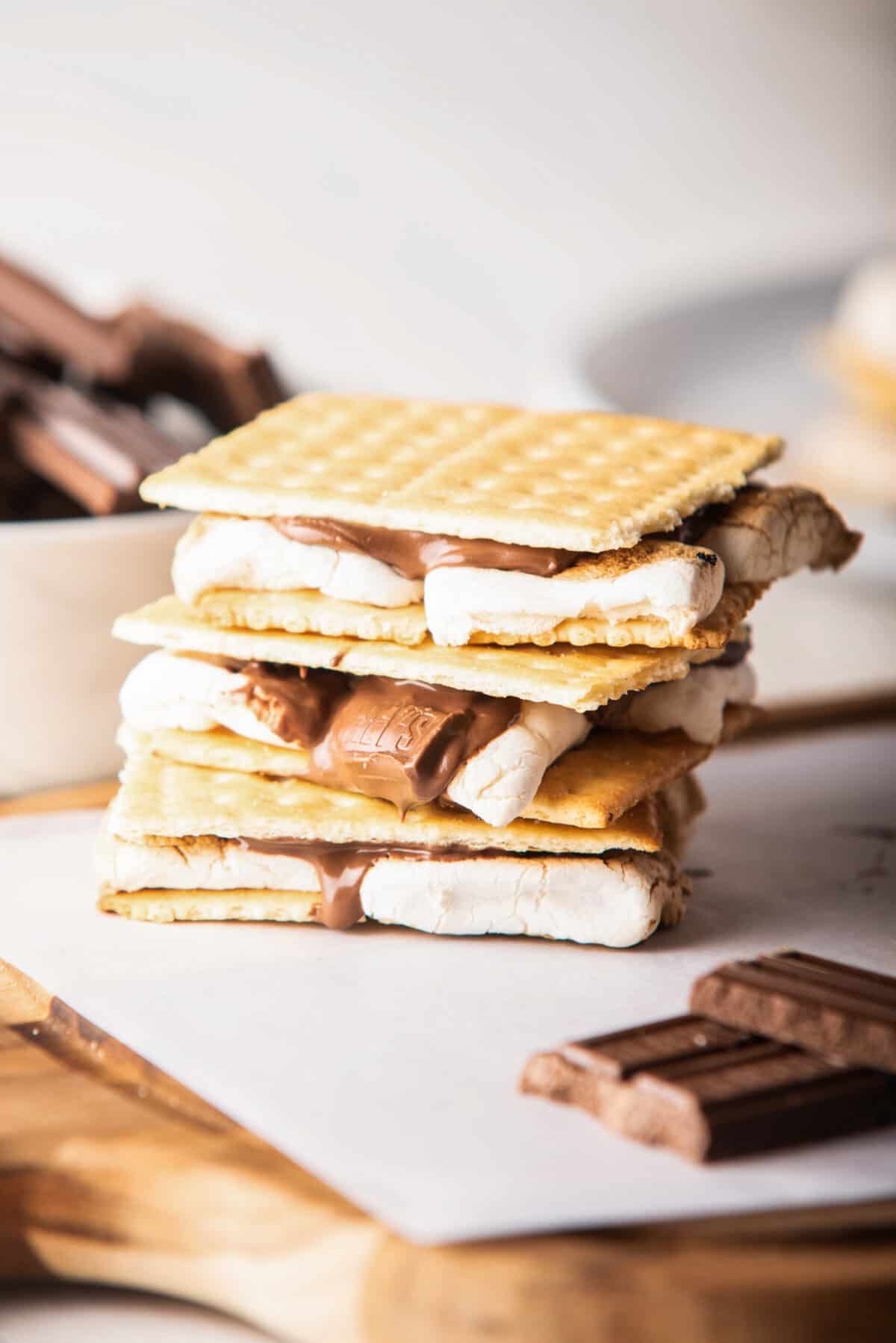 A straight view of cooked air fryer s'mores above a wooden board next to pieces of chocolate.