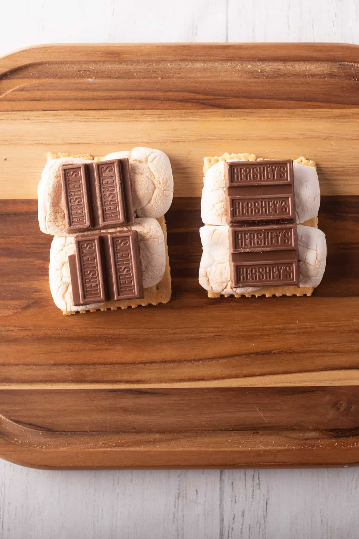 Overhead view of two graham crackers with marshmallows and chocolate pieces on top, placed above a wooden board.