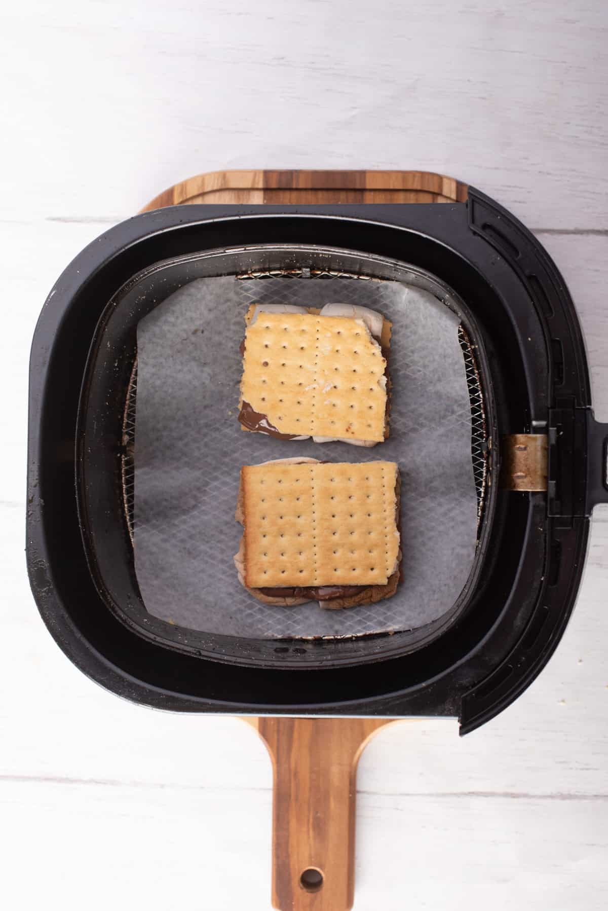 Overhead view of two Graham crackers filled with chocolate and marshmallows placed on an air fryer basket.