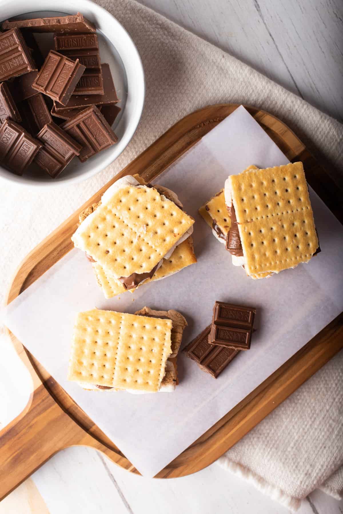Overhead view of three air-fried s'mores placed on a wooden board next to a bowl of chocolate pieces.