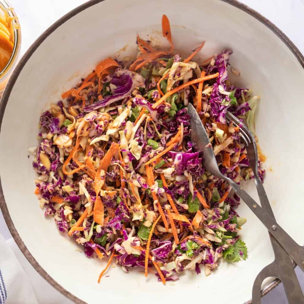 Mixed slaw in a large bowl with an empty bowl of peanut dressing on the side (with tongs)