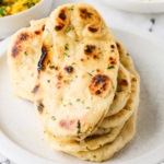 Stack of homemade garlic naan on a white plate with curry and rice in the background.