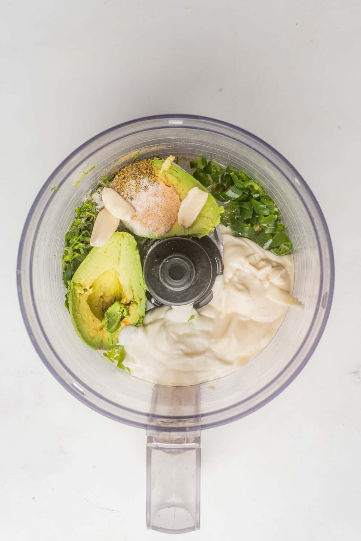 An overhead image of avocado, cilantro, garlic, mayonnaise, sour cream, lime juice, and seasoning, placed in a blender.