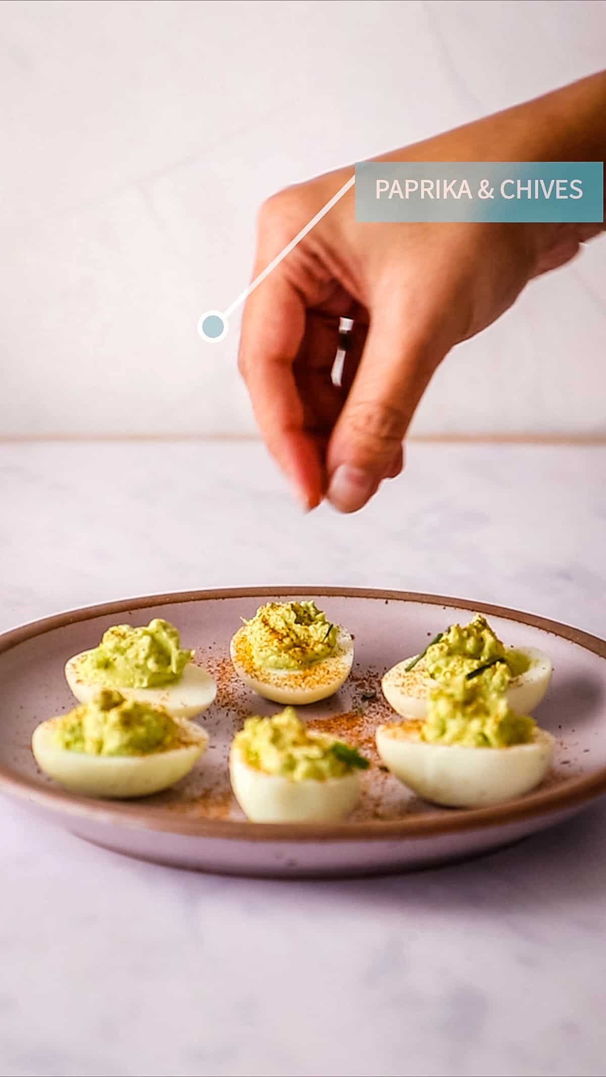 Hand sprinkling paprika over finished deviled eggs in lilac plate.