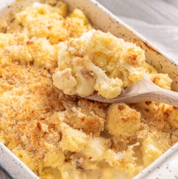 An image of baked cauliflower with cheese in a baking dish, with a full serving spoon resting on top of it,