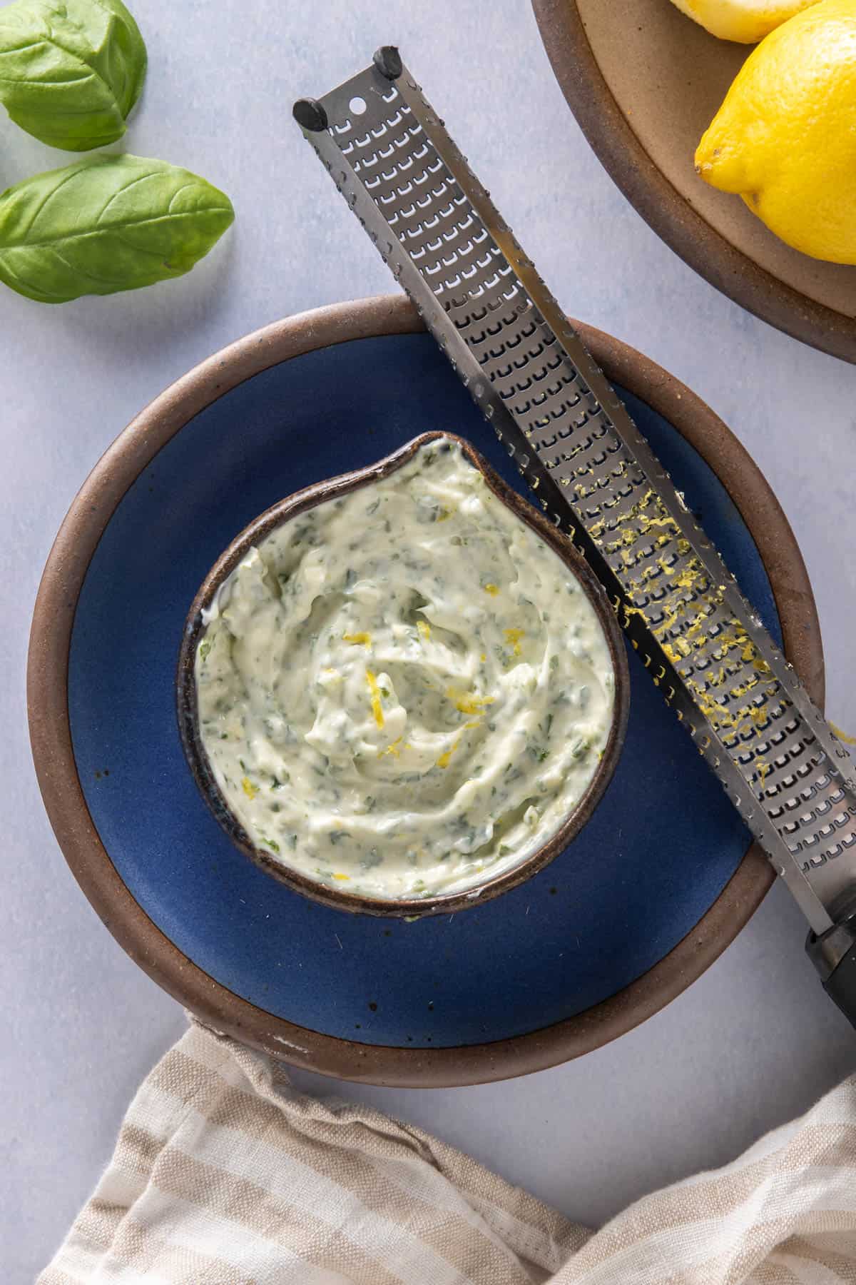 Basil aioli in a jar, on a blue plate, with a microplane with lemon zest resting on it. Basil, lemons in background