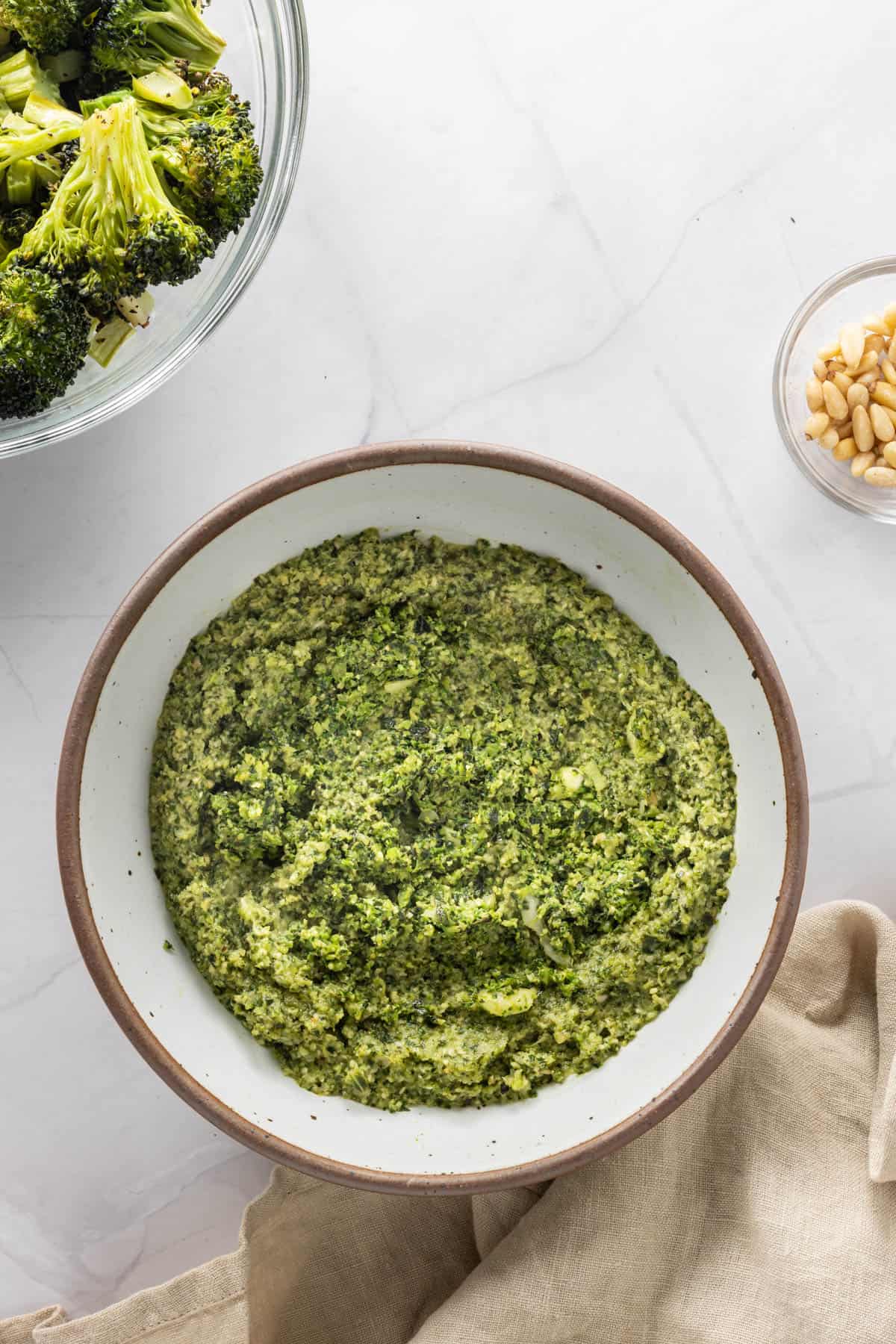 Bowl of broccoli pesto with extra broccoli and pine nuts in background