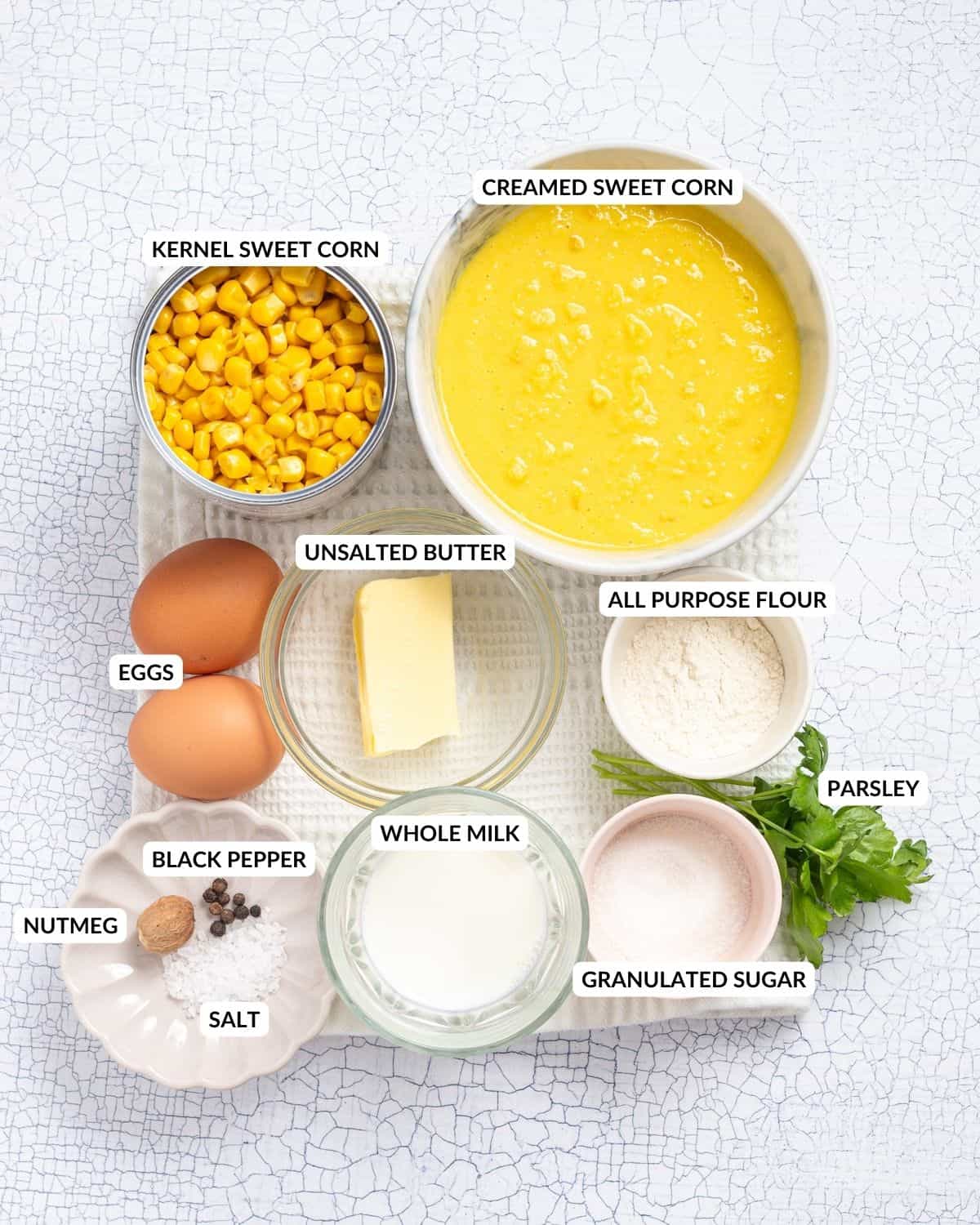 The ingredients of corn casserole arranged side by side with labels. It includes sweet corn kernels, creamed corn, two brown eggs, butter, whole milk, AP flour, sugar, parsley, and seasonings.