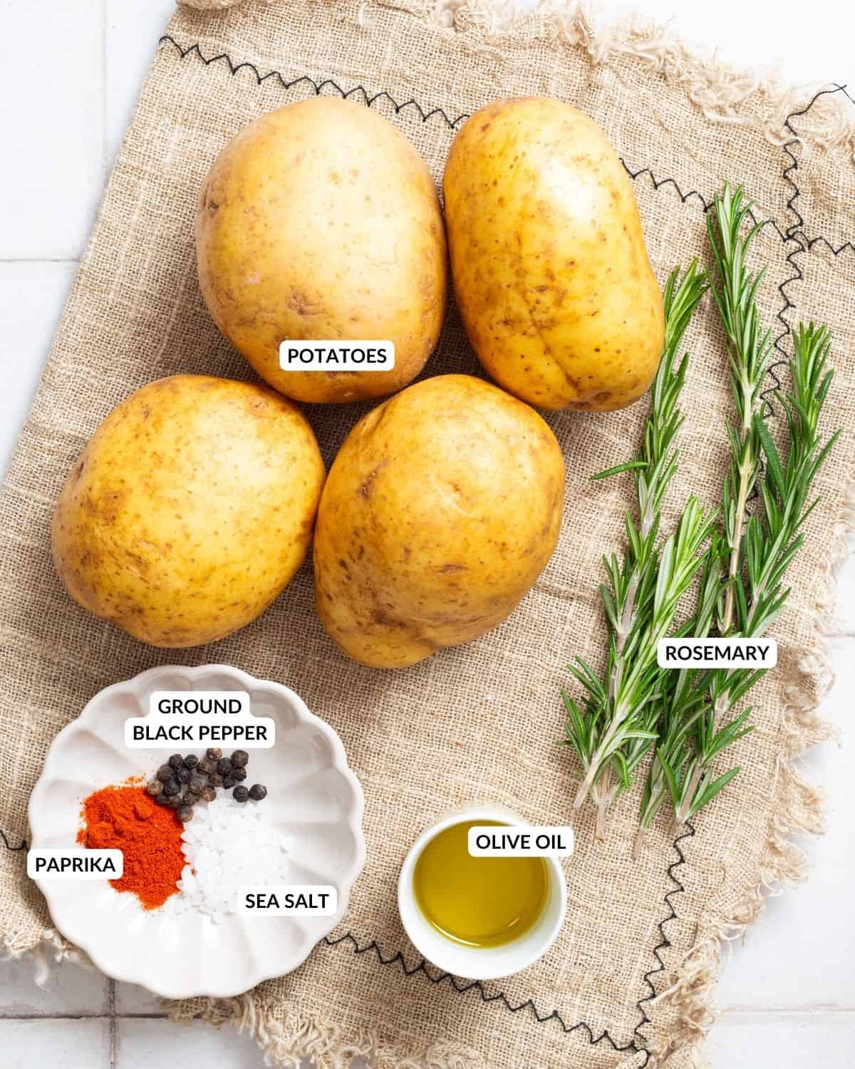 An overhead image of four russet potatoes, a spice mixture on a small plate, olive oil in a small container, and fresh rosemary twigs, with labels.