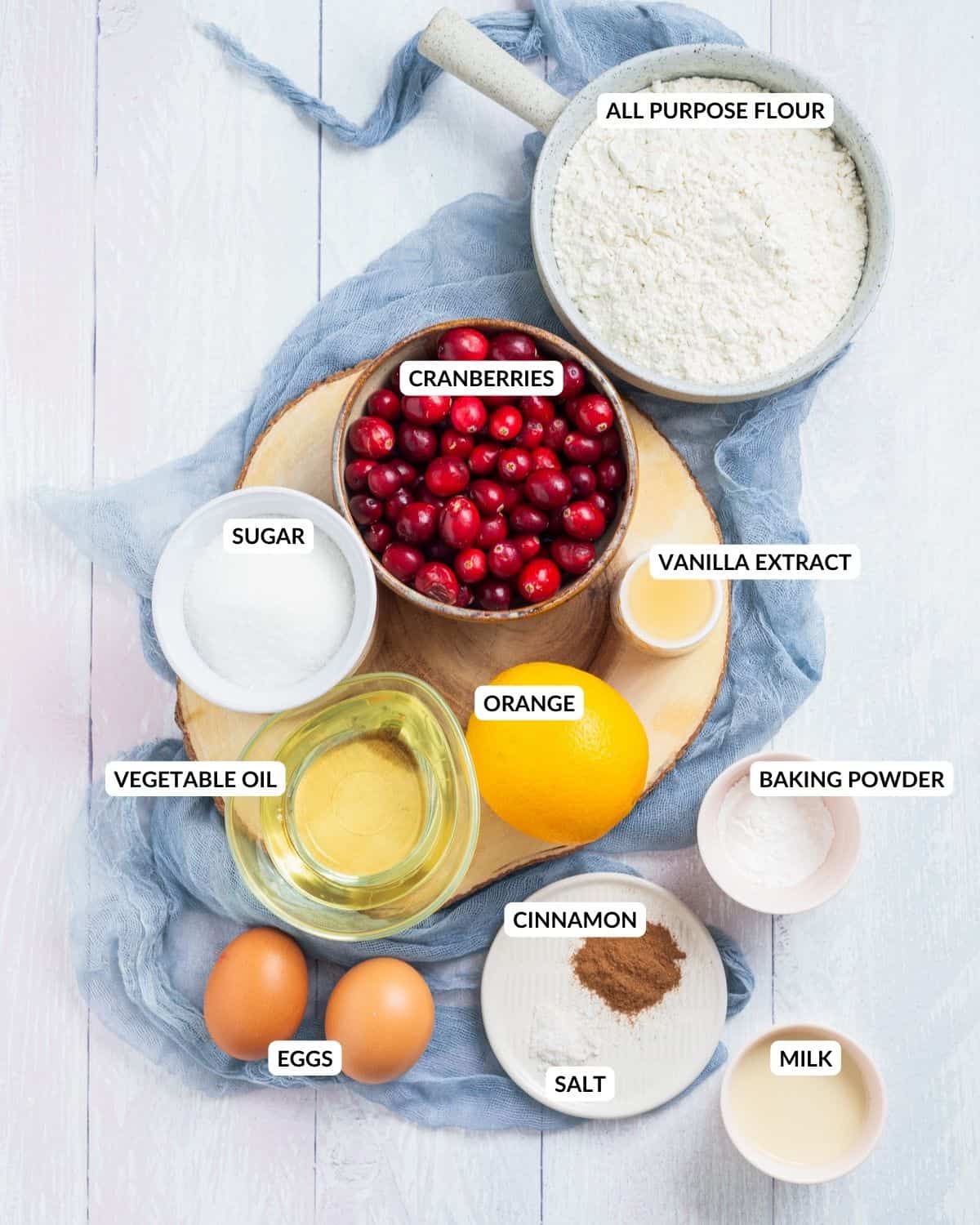An overhead image of cranberries, flour, baking powder, eggs, vanilla extract, an orange, milk, sugar, cinnamon, and salt in separate containers and a label for each ingredient.