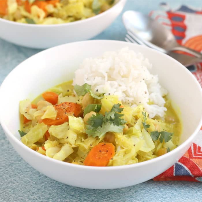 An straight view of cabbage and chickpea coconut curry placed in a white bowl with a scoop of rice on top and a spoon and fork on the side.
