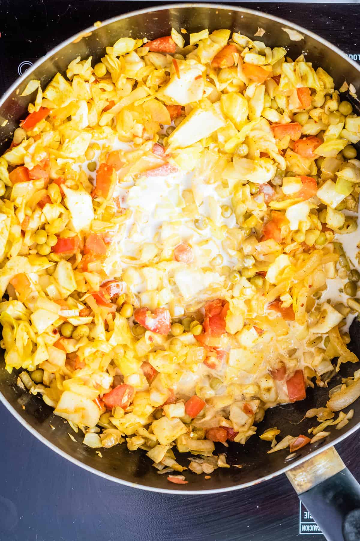 Overhead view of mixed chopped onions, garlic, ginger, tomatoes, peas, turmeric, garam masala, red chili powder, and chopped cabbage added with coconut milk.