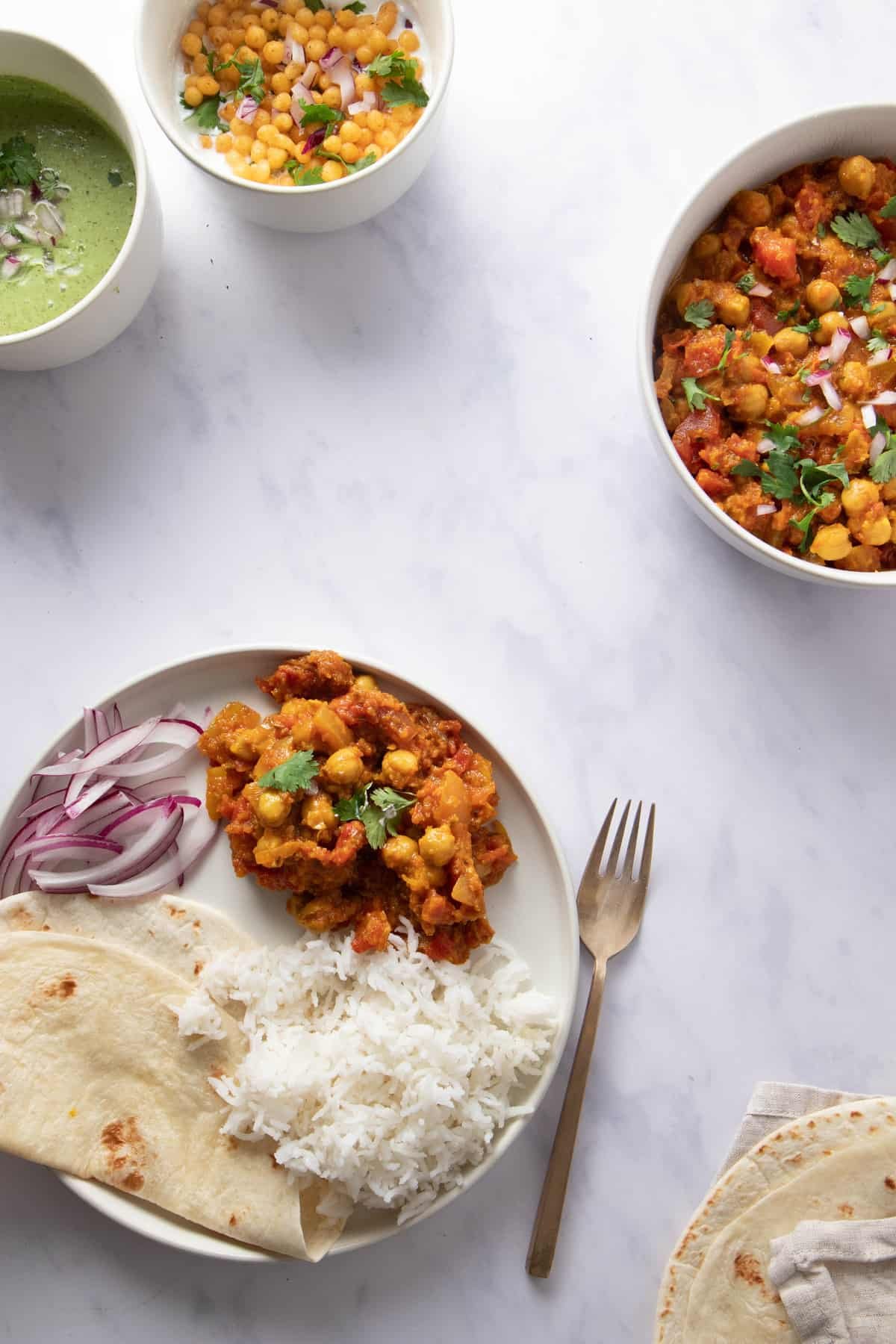 Plate with chana masala, rice and flatbread as well as a bowl of chana masala and raita in the background