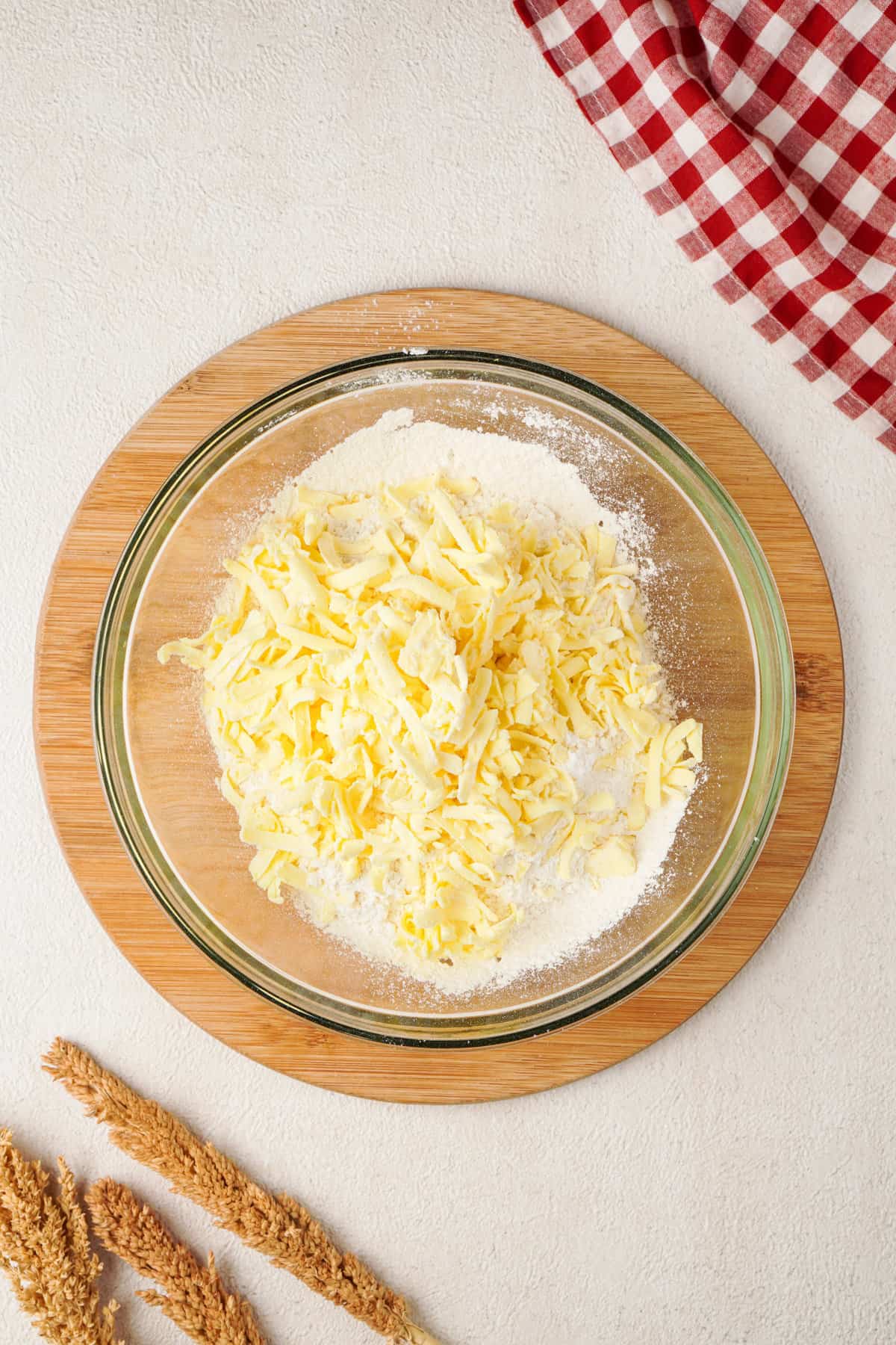 Overhead view of shredded butter added to glass bowl with dry ingredients, placed on top of wooden platter. 