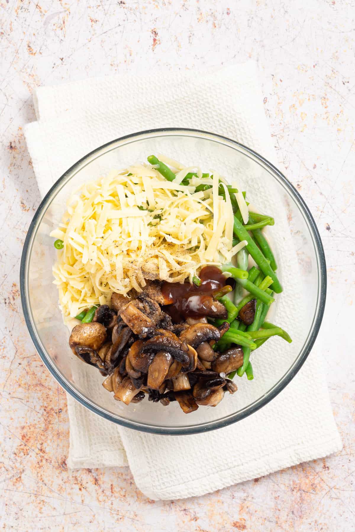 An overhead image of cooked mushrooms, blanched green beans, and cheddar cheese in a clear bowl.