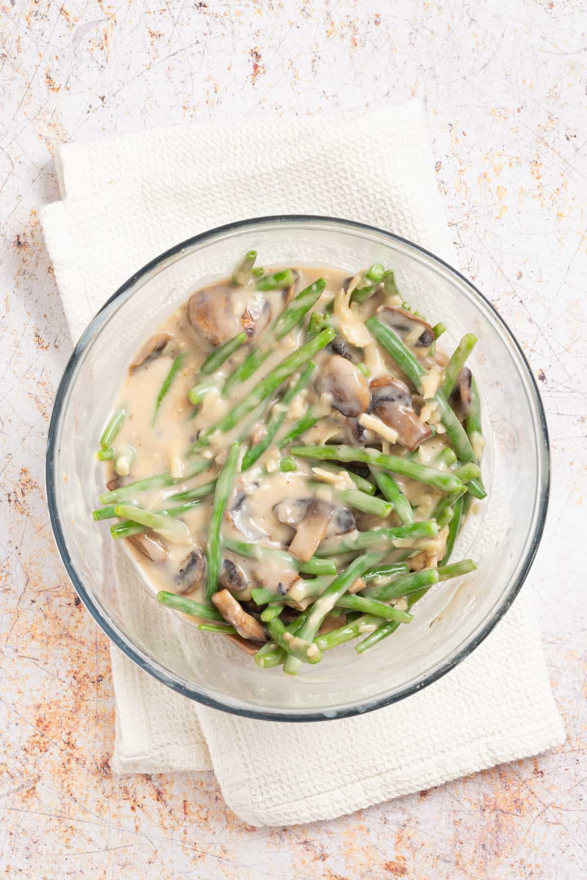 An overhead image of condensed mushroom soup, cheddar cheese, Worcestershire sauce, milk, salt and black pepper added to a clear bowl with green beans, mushrooms, and cheddar cheese.