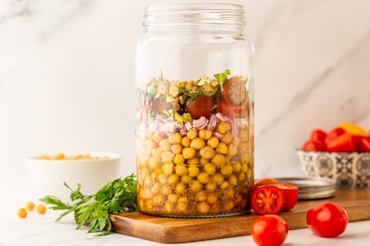 Layered chickpea salad in a mason jar with tomatoes, herbs, shallots, and pistachios stacked above the lemon vinaigrette dressing.