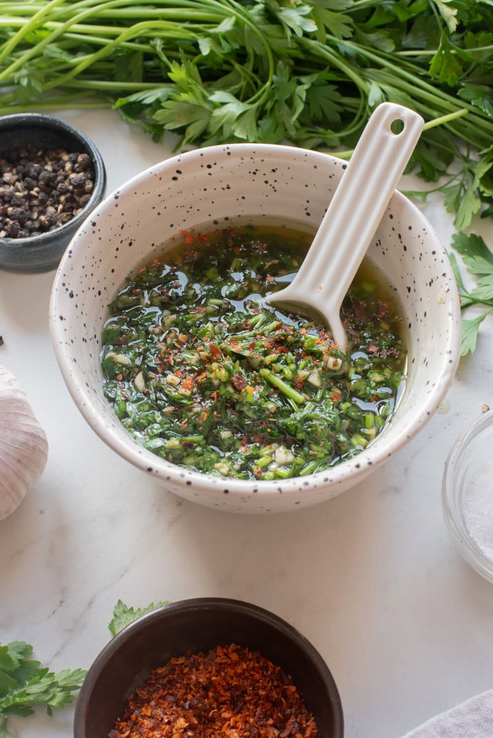 Spoon lifting fresh chimichurri sauce from a bowl with visible parsley, garlic, olive oil, and red pepper flakes.