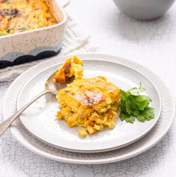 An image of a single serving of corn casserole on a plate with a spoon beside it and the casserole behind it.