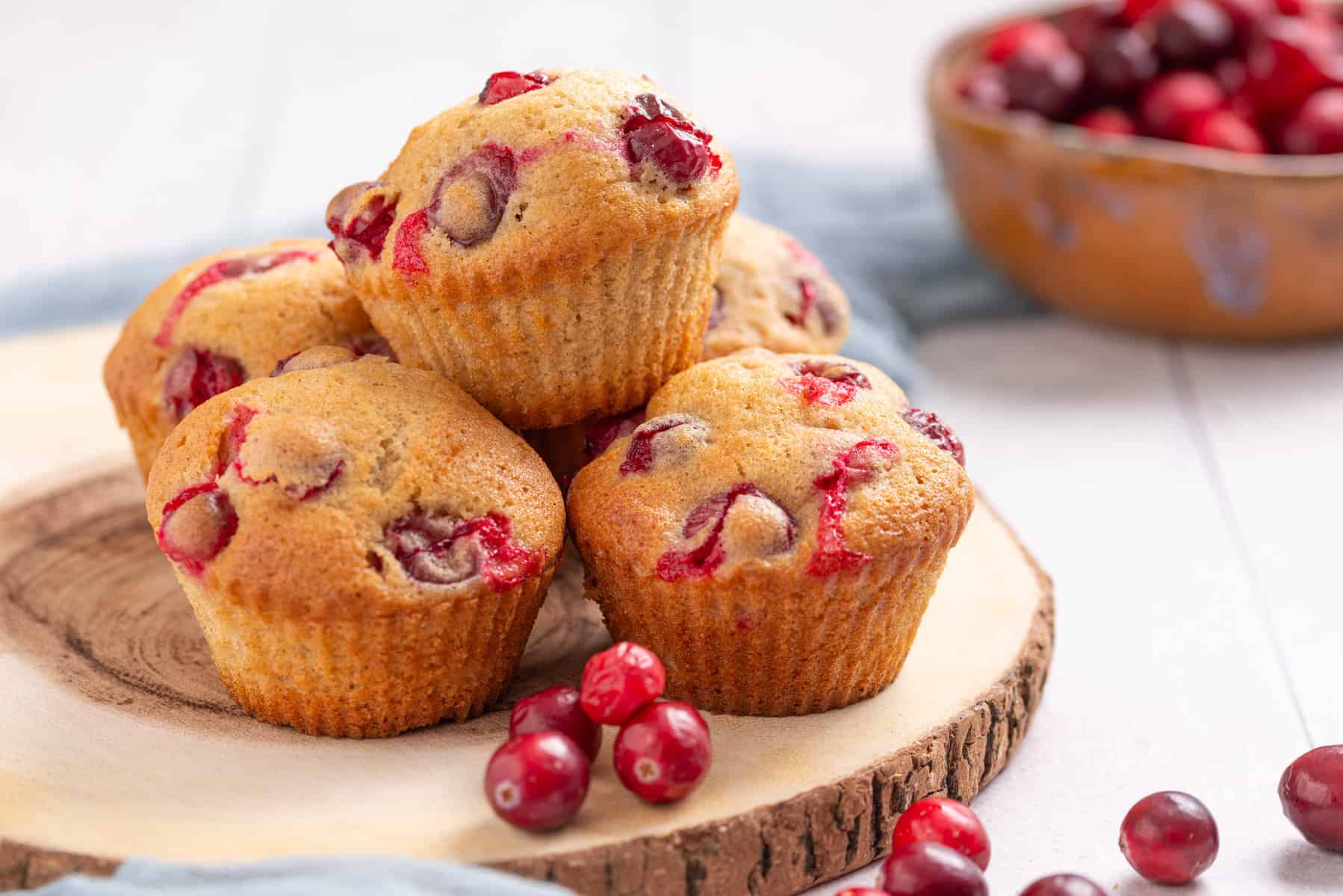 An image of a batch of cranberry orange muffins, one stacked on top of the others, on a wooden serving board.