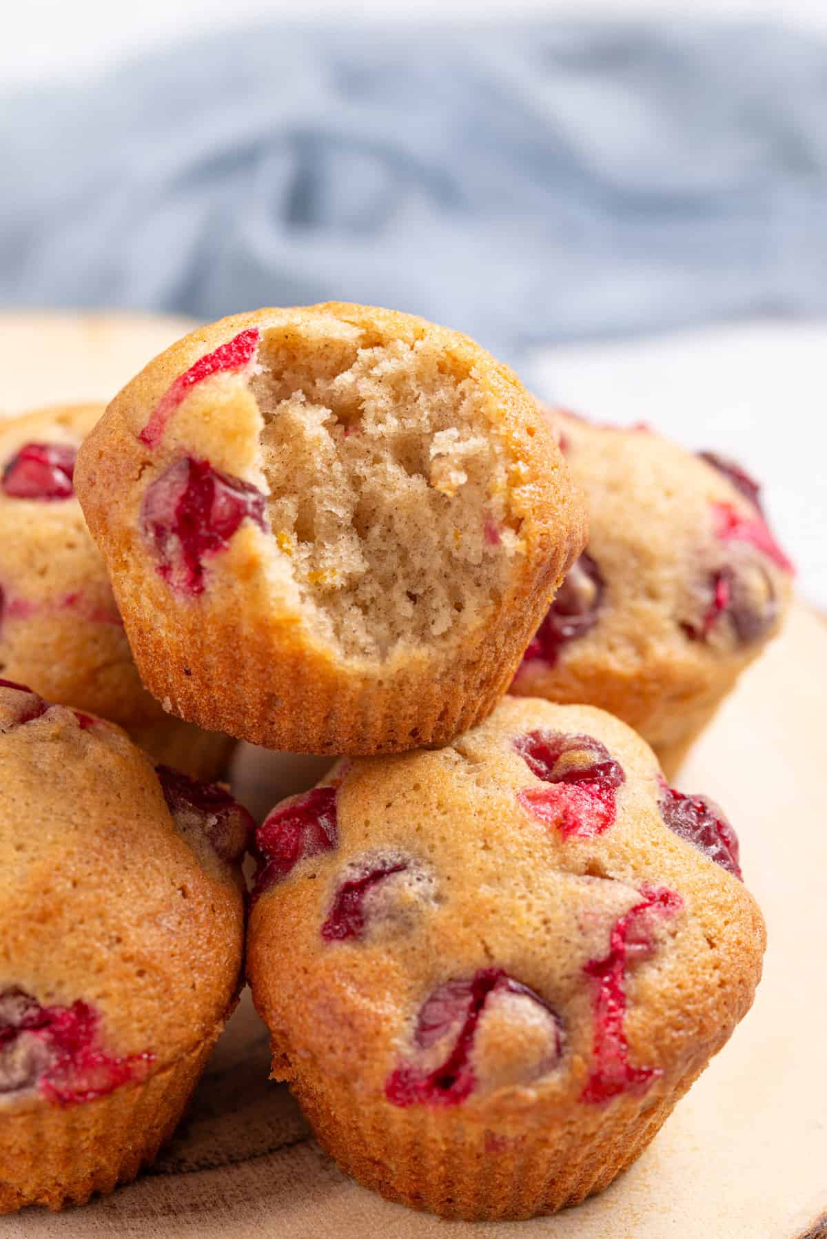 An image of a batch of cranberry orange muffins, one muffin with a bite stacked on top of the others, on a wooden serving board.
