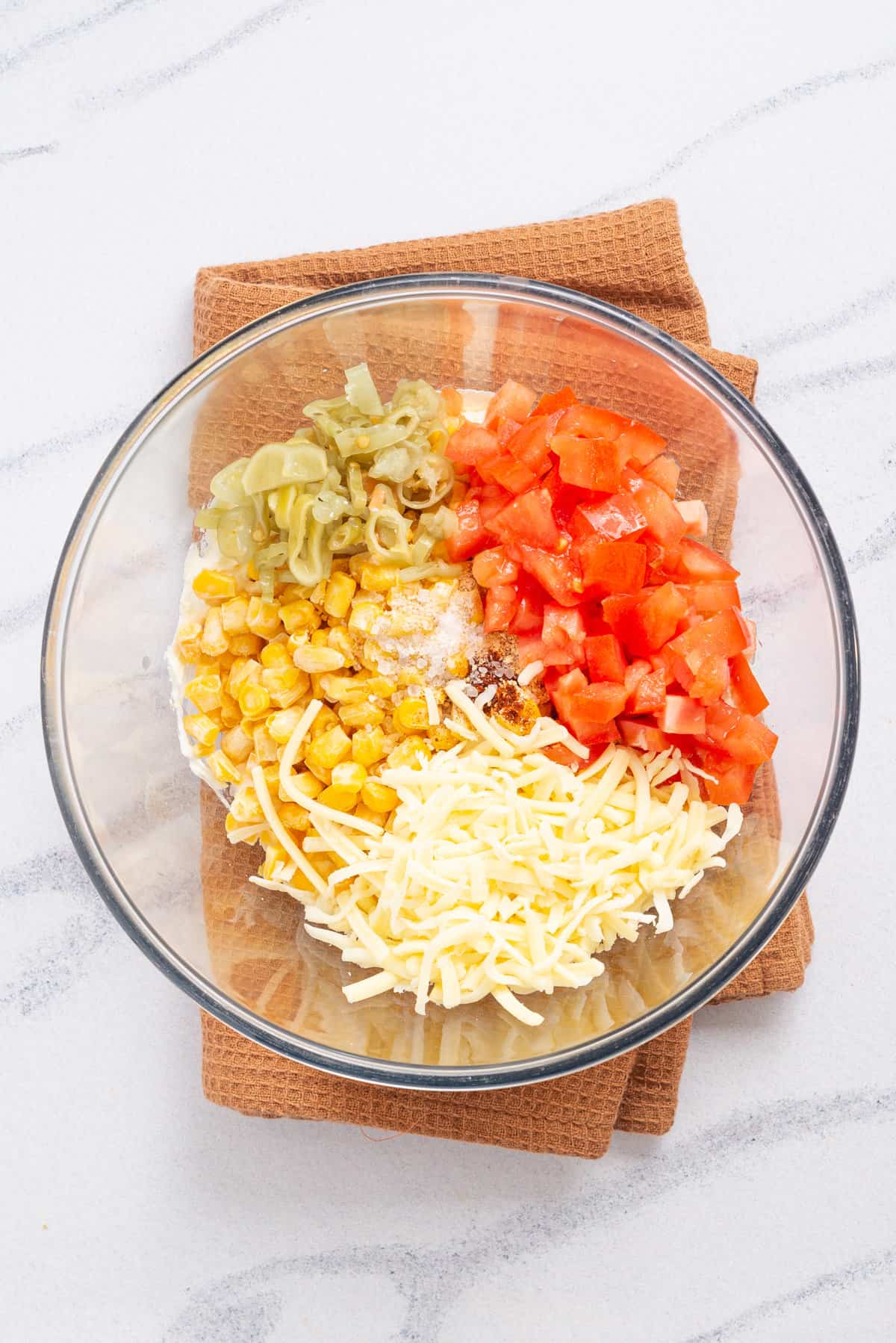 An overhead image of a bowl with sweet corn, onion powder, chili powder, salt, pepperoncinis, tomatoes, cheddar cheese before mixing together.
