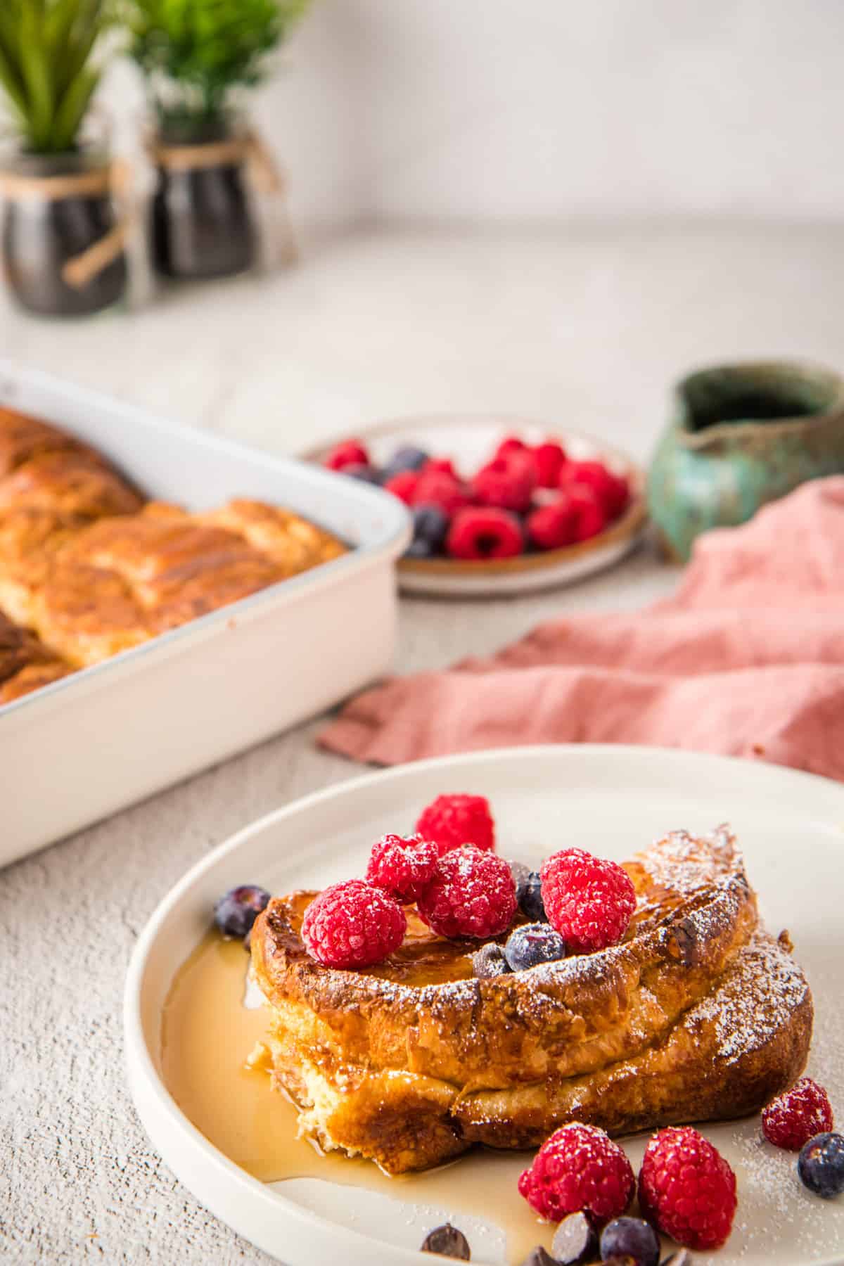 Close up of finished dish on white plate, with berries on top, and baking tray with croissant French toast bake in the background.