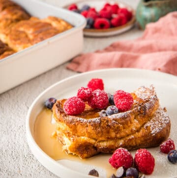 Close up of croissant French toast bake slices on white plate, with berries and maple syrup on top