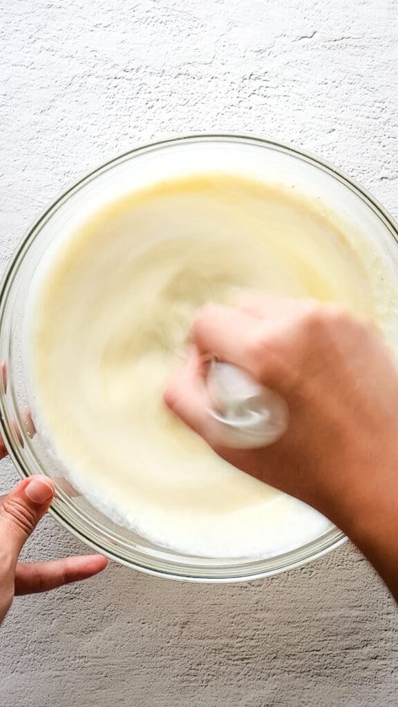 Overhead view of custard fully mixed, with hand and whisk. 