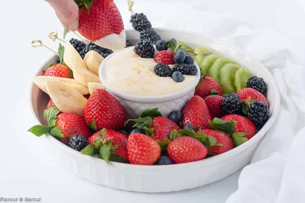 A straight view of a hand dipping a strawberry into creamy lemon curd fruit dip.