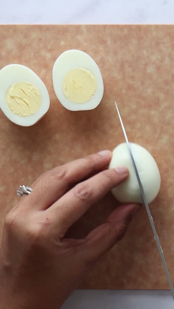 Slicing into an egg lengthwise. Two cut egg halves in background on composite cutting board.