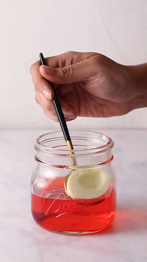 Placing egg white half into glass jar with colored water using a black and gold spoon.