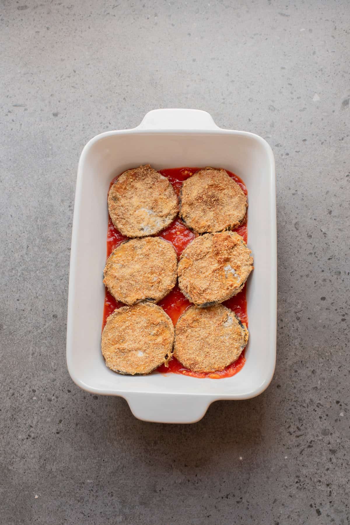 An overhead image is layering eggplant slices, marinara sauce, and cheese in a baking sheet.