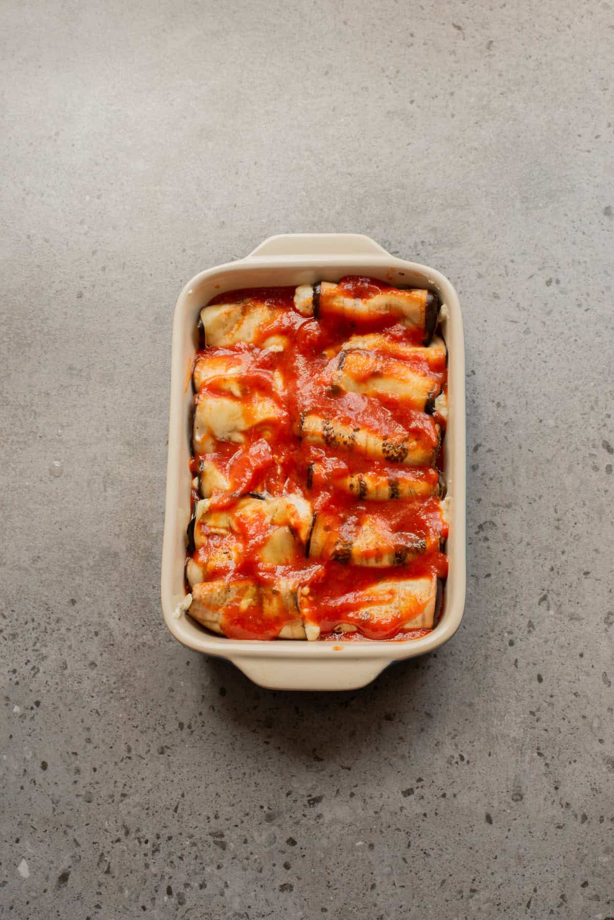 An overhead image of spooning marinara sauce over the eggplant slices in a baking dish.