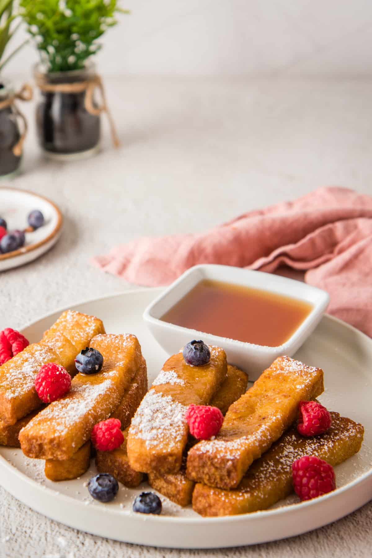 Straight on view of French toast sticks on a white plate, with berries on top and maple syrup in small container on plate.