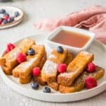 Close up of French Toast Sticks with berries on top on a white plate, with maple syrup on the side. More berries in background.