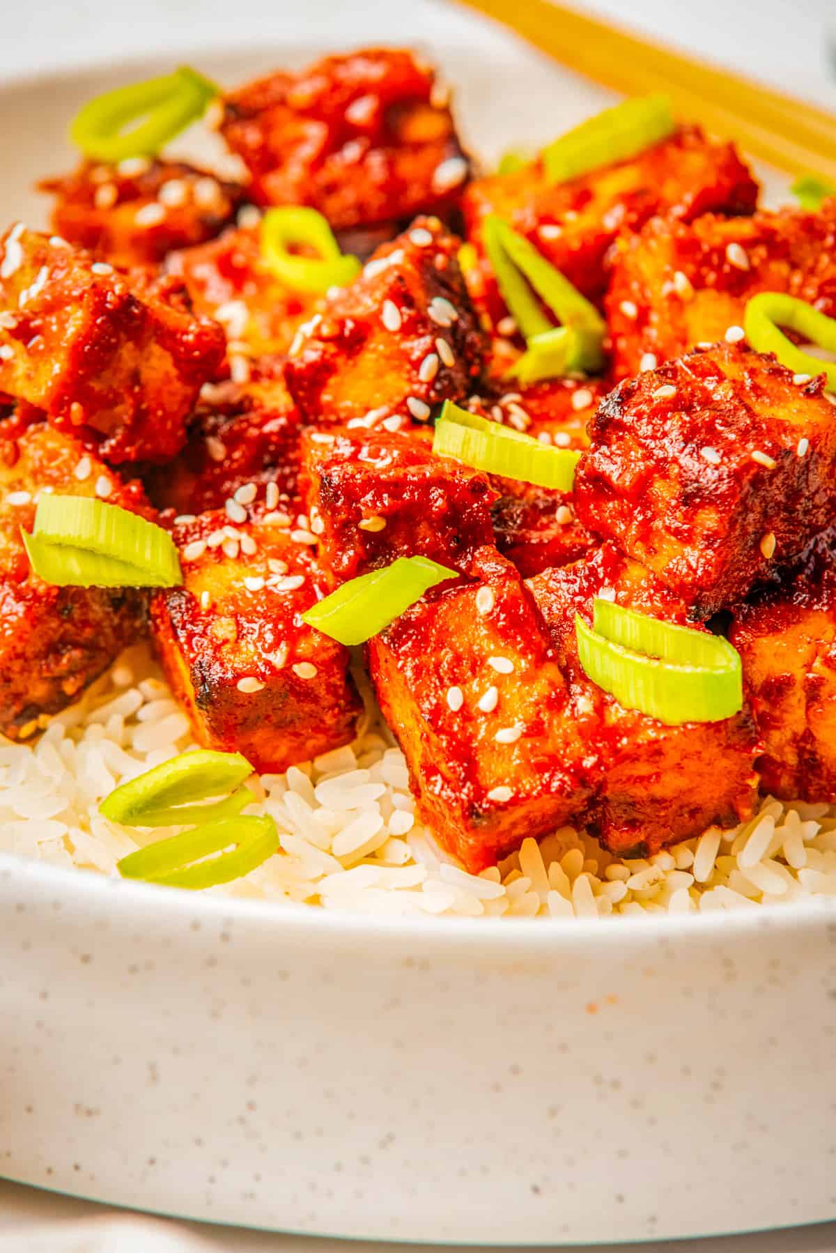 A close up image of gochujang tofu with sesame seeds and green onions, on top of a bowl of rice.