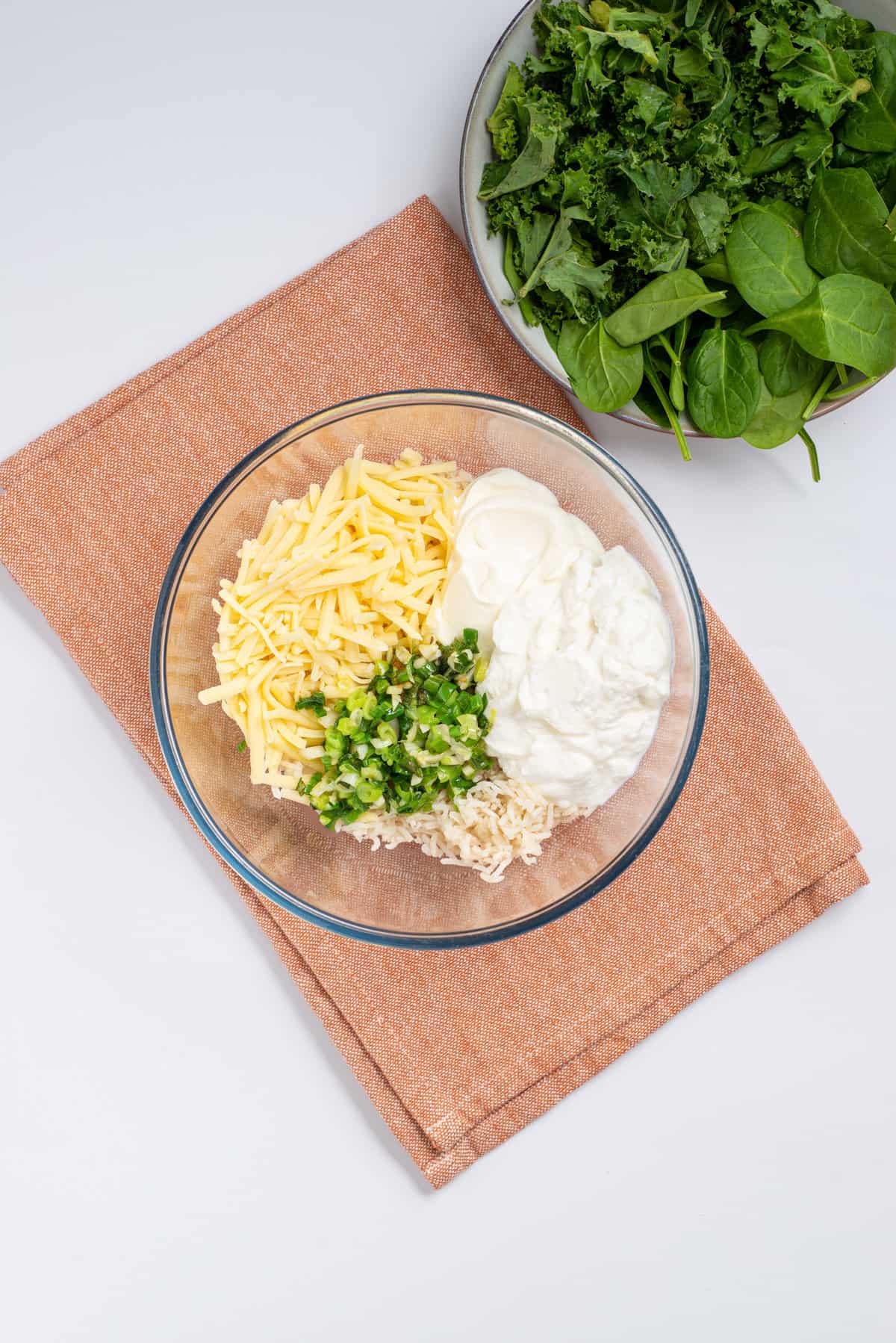 Overhead view of a transparent bowl with cooked rice, cheddar cheese, ricotta, sour cream, spinach, kale, sauteed garlic, scallions, and chiles. 