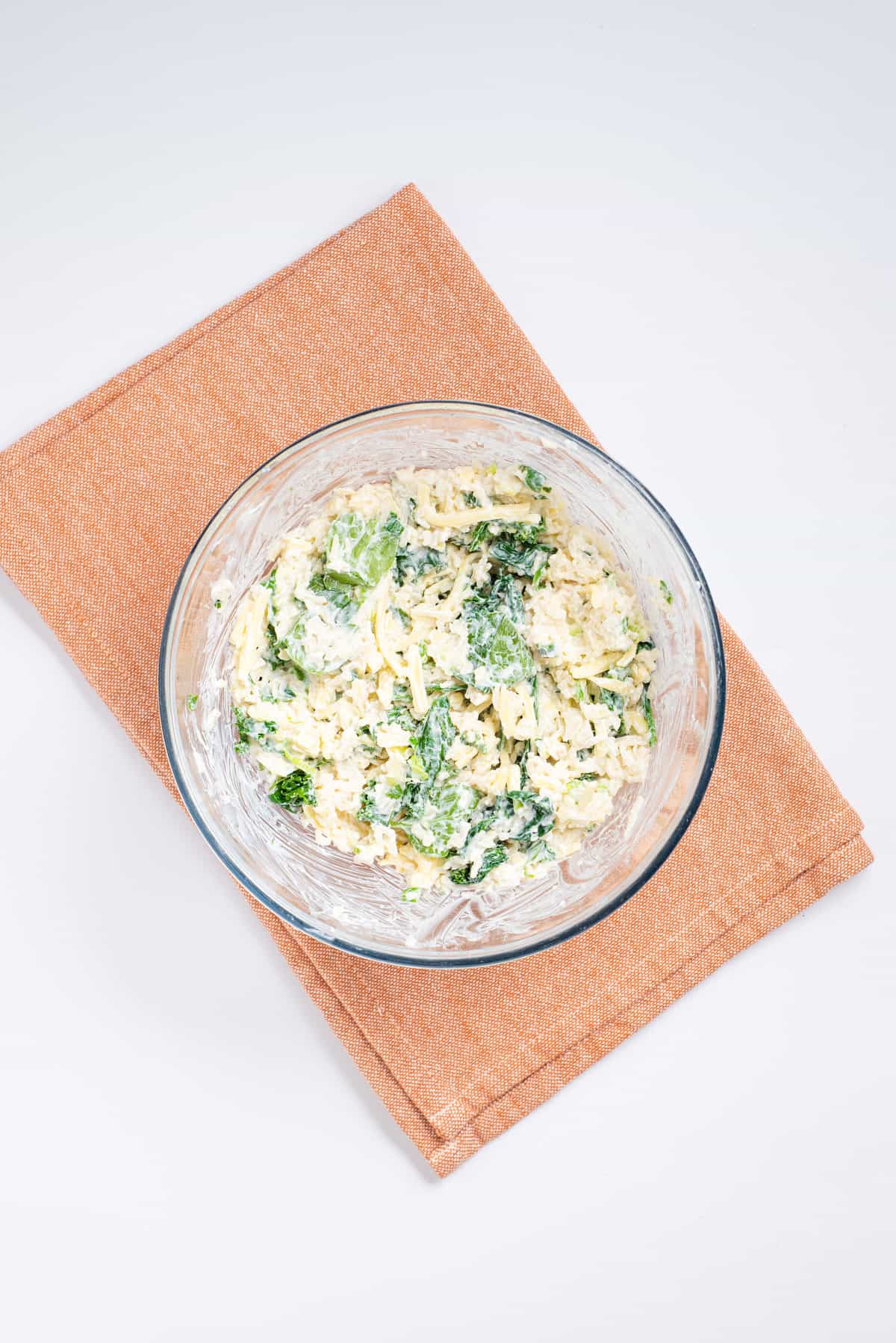 Overhead view of a transparent bowl with mixed cooked rice, cheddar cheese, ricotta, sour cream, spinach, kale, sauteed garlic, scallions, and chiles. 