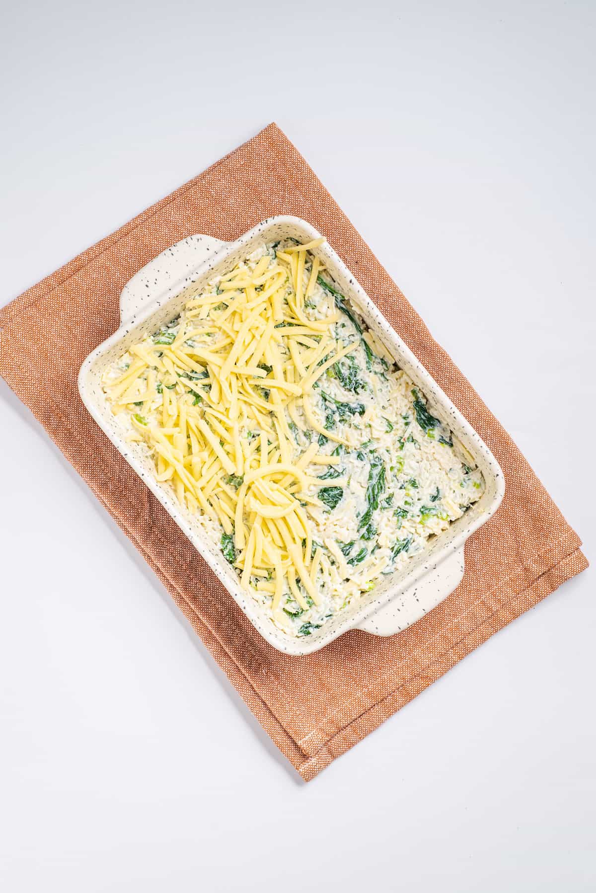 Overhead view of an uncooked green rice casserole topped with cheese on a white baking dish. 