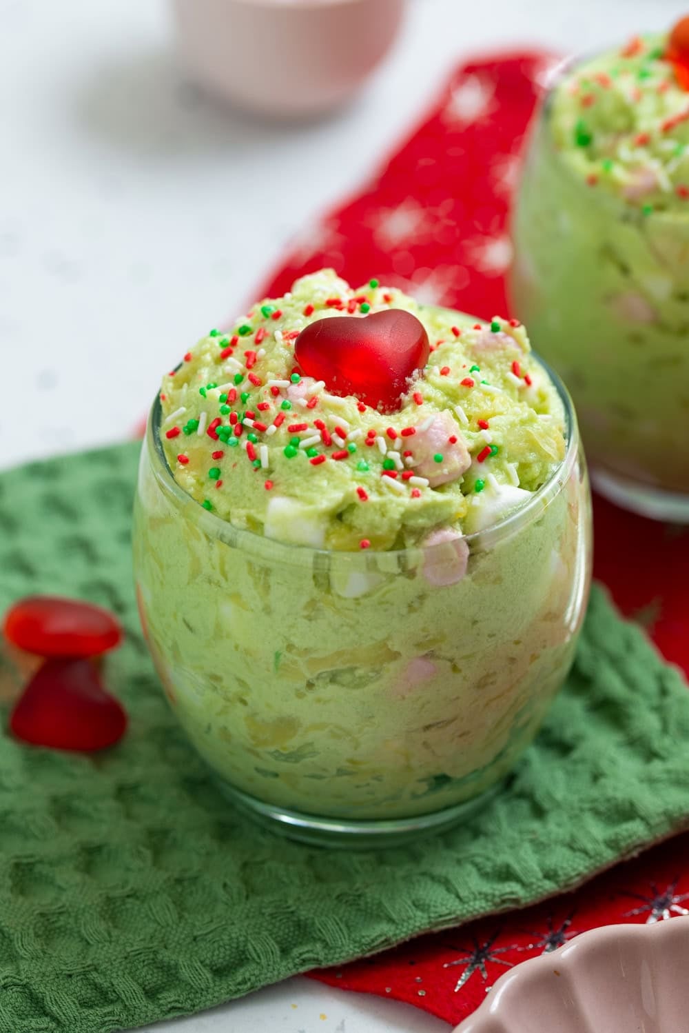 Finished green Grinch fluff topped with a red candy heart and red and green Christmas sprinkles in a clear glass.