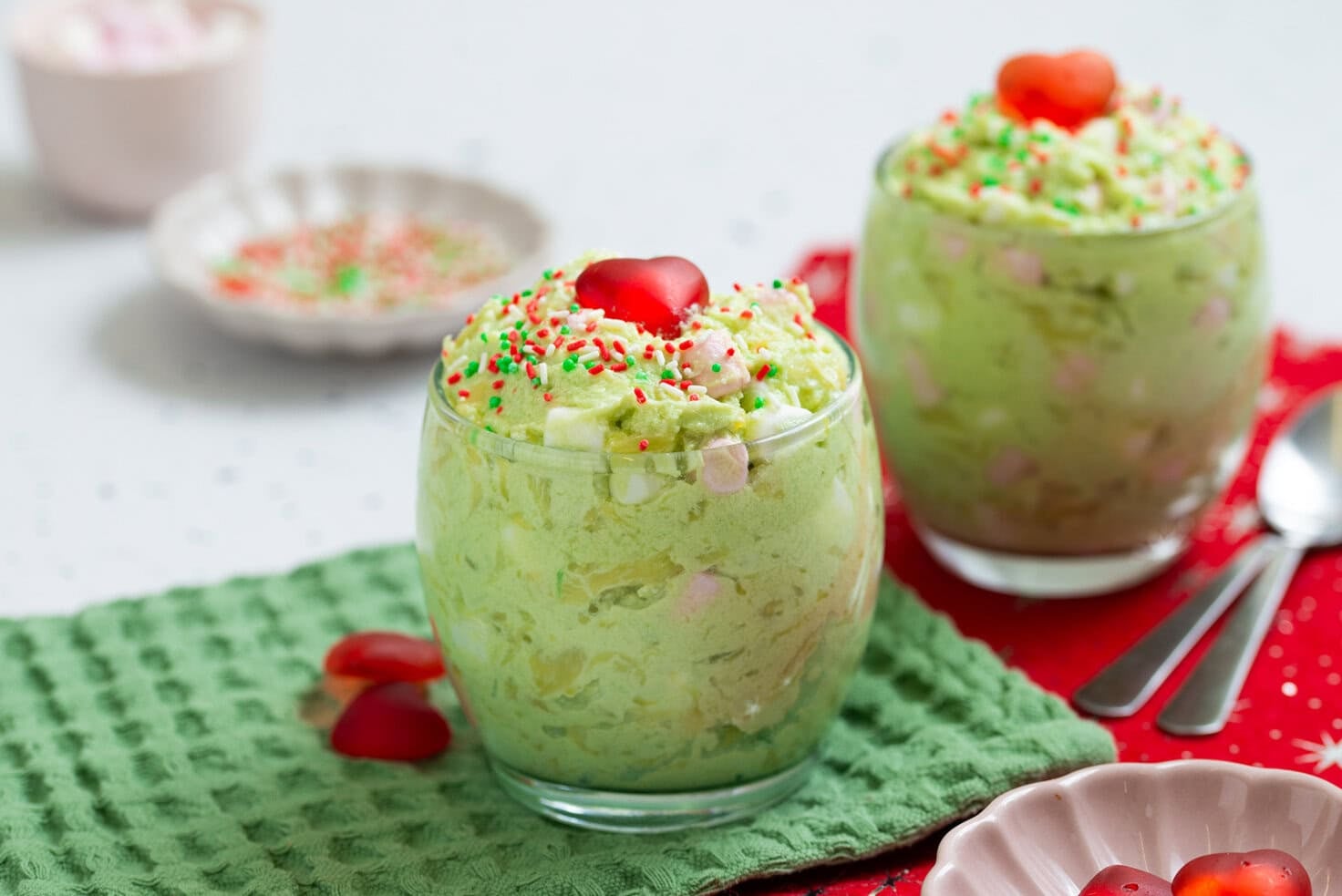 Two glasses of green Grinch fluff topped with red candy hearts and Christmas sprinkles on a green napkin.