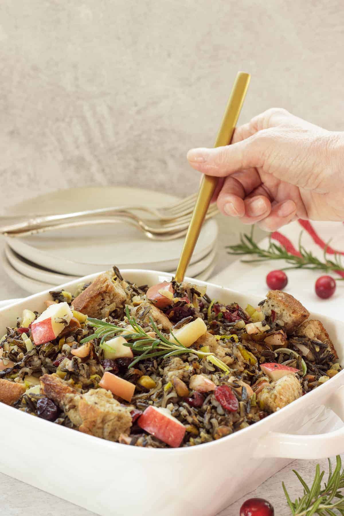 A straight view of wild rice stuffing placed on a white casserole with a hand scooping the dish out using a golden spoon.