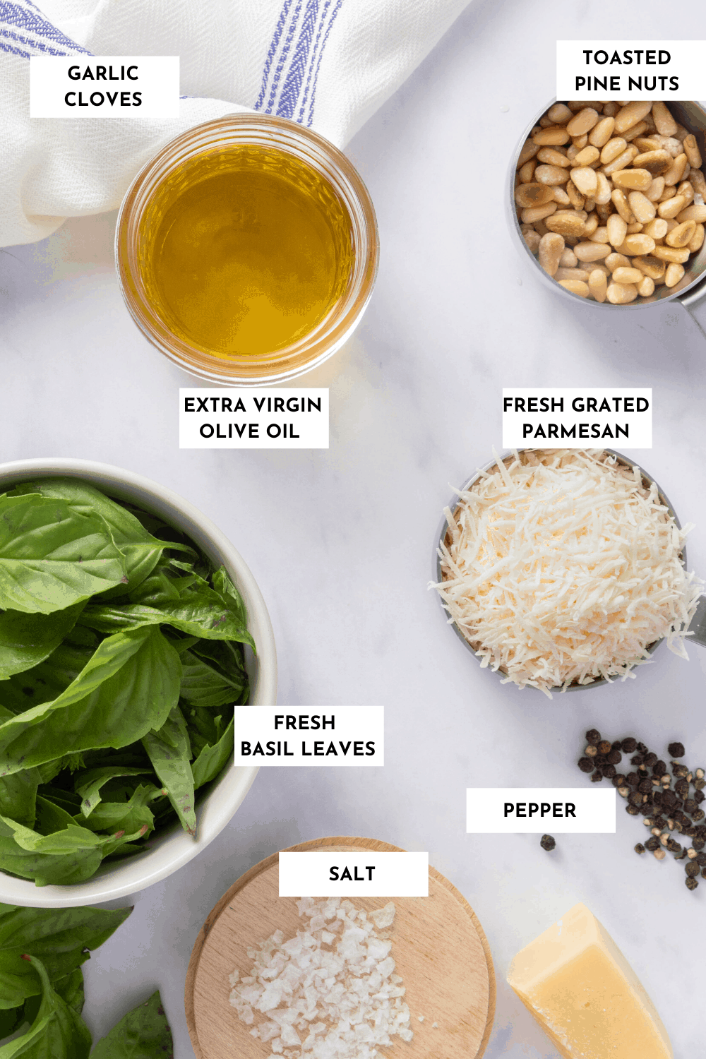 Ingredients for making basil pesto, labeled on a table - basil, olive oil, salt, pepper, parmesan and garlic (not pictured)