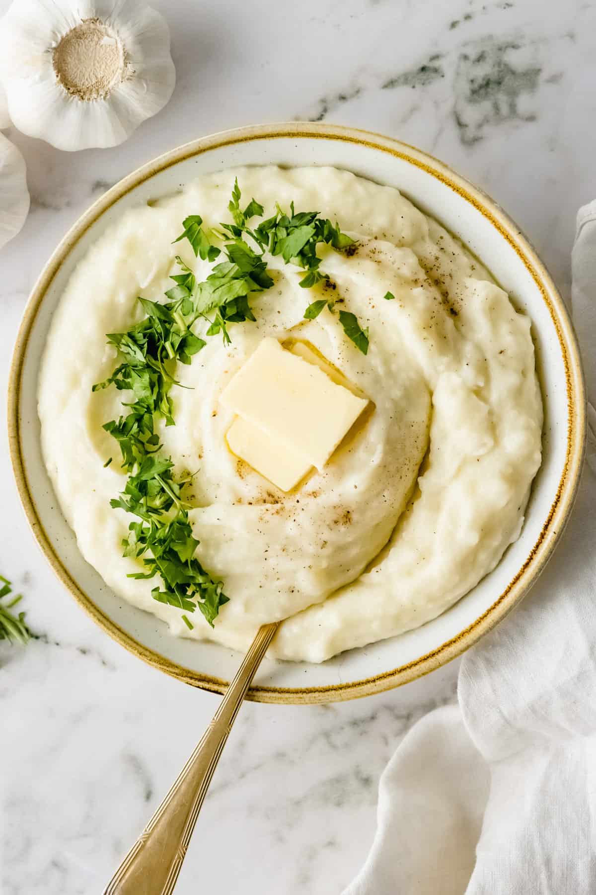 Close up of mashed potatoes in a white bowl with a spot of butter and some herbs on top, golden spoon in it.